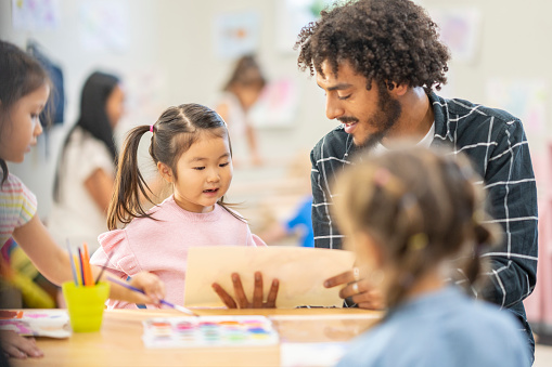 A male childcare worker holds out a child's artwork as he takes a closer look. b The little girl of Asian decent is sitting beside him as she looks up for his approval.