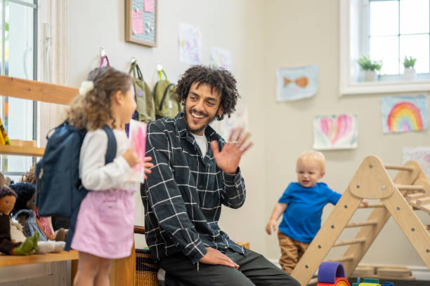 Leaving Daycare A sweet little girl packs up her things and places her backpack on as she prepares to leave at the end of the day. A male childcare worker is waving goodbye to the little girl as she heads out. preschool classroom jobs stock pictures, royalty-free photos & images