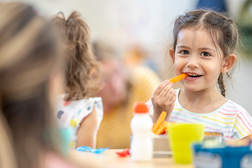 Daycare children are seen sitting around a table eating their lunch.  They are dressed casually and have healthy eating choices on the table in front of them as they talk amongst themselves.