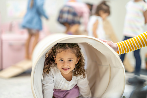 A sweet little girl crawls through a fabric tunnel at daycare as she explores the room with her friends.  She is dressed casually and smiling as she crawls through the tunnel.