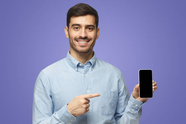 istock Man in blue shirt on purple background, presenting smartphone pointing at blank screen 1431057972