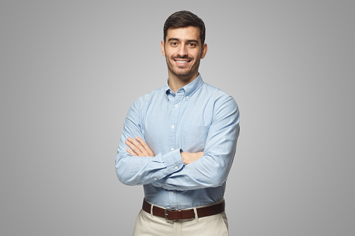 Handsome smiling business man in blue shirt standing with crossed arms, isolated on gray background