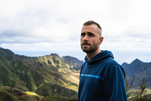 Caucasian young attractive man with blue hoodie and cloudy day, looking serious and observing the landscape