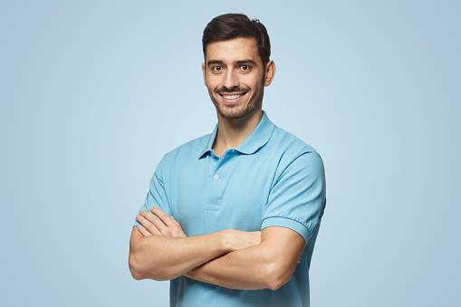 Portrait of attractive young sporty man in blue polo shirt standing with crossed arms, isolated on blue background