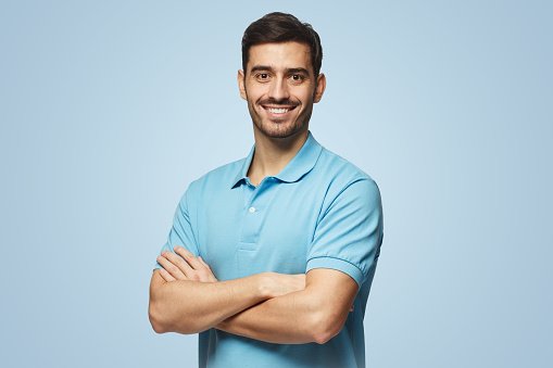 Portrait of young european caucasian man isolated on blue background, standing in blue polo shirt with crossed arms, smiling and looking at camera
