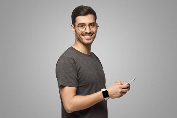 istock Young man standing isolated on gray background, holding phone, looking at camera with smile 1431059414