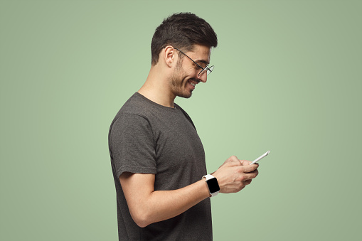 Side portrait of handsome man looking at screen of smartphone while exchanging messages, isolated on green background