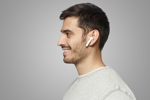 Sideways portrait of smiling young man listening to music or radio, uses modern wireless earphones
