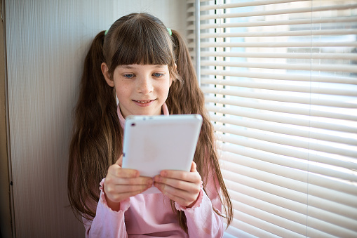 a little girl with freckles and blue eyes sitting by the window and using a tablet.