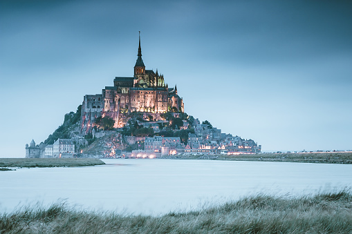 France, Normandy, Manche department, Bay of Mont Saint-Michel Unesco World Heritage, Abbey of Mont Saint-Michel. Long exposure sunset view of Le Mont Saint-Michael with mistyc aural and deep blue waters in summer evening light at sunset in summer, Normandy, northern France