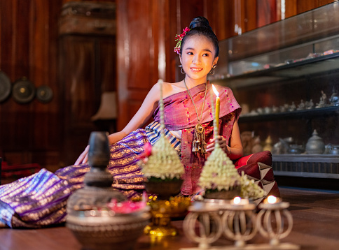 Young beautiful asian Laos lady wearing traditional Lao costume dresses sitting with an elegant pose with some flower bouquet and candles prepared for Loy Krathong festival in front of her