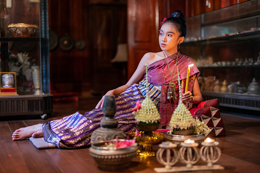 Young beautiful asian Laos lady wearing traditional Lao costume dresses sitting with an elegant pose with some flower bouquet and candles prepared for Loy Krathong festival in front of her