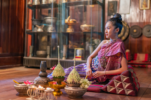 Young beautiful asian Laos lady wearing traditional Lao costume dresses sitting with an elegant pose with some flower bouquet and candles prepared for Loy Krathong festival in front of her