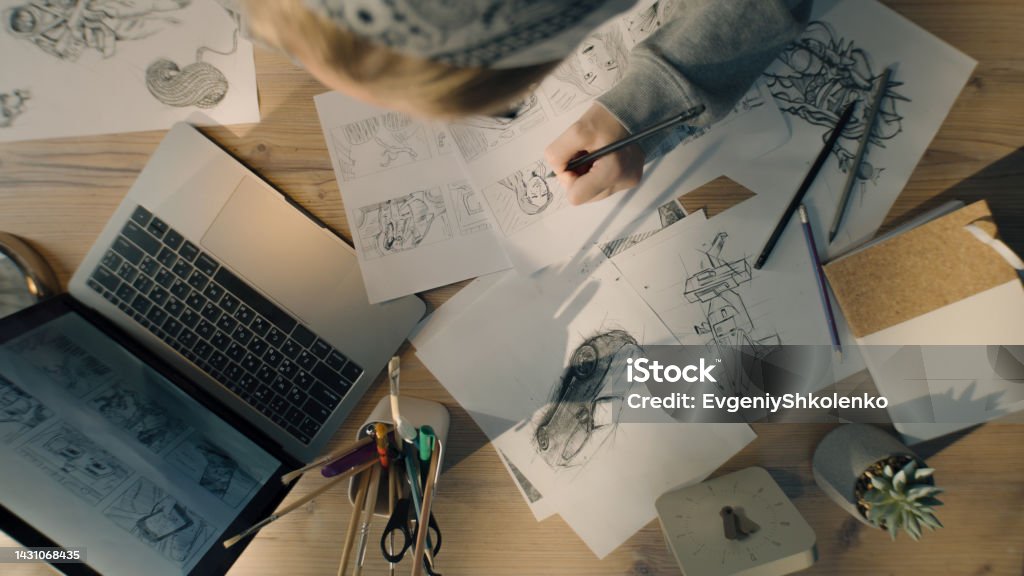 Young woman artist draws pencil sketches works on storyboard of a comic top view Top view of young woman drawing pencil sketches. Working on a storyboard of a comic in a design studio. Designer work table with a laptop and stationary jar. Animator Stock Photo