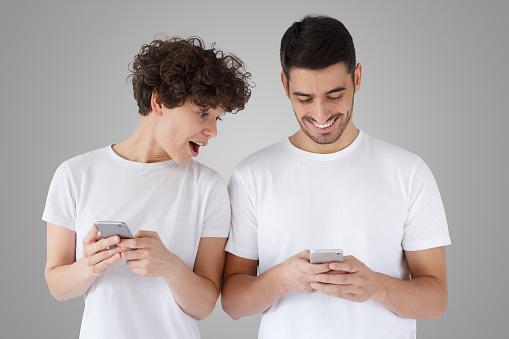 Surprised young woman looking at smartphone screen of her boyfriend with mouth open, isolated on gray background