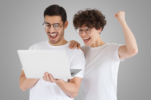 Happy excited couple holding laptop and raising arms up to celebrate achievement, isolated on gray background