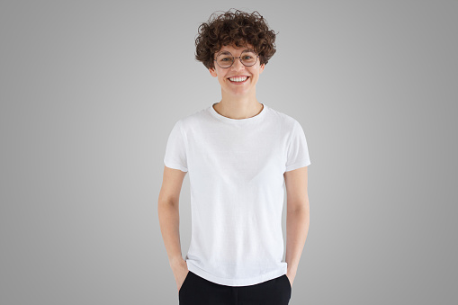 Young european woman standing with hands in pockets, wearing blank white t-shirt with copy space for your logo or text, isolated on gray background