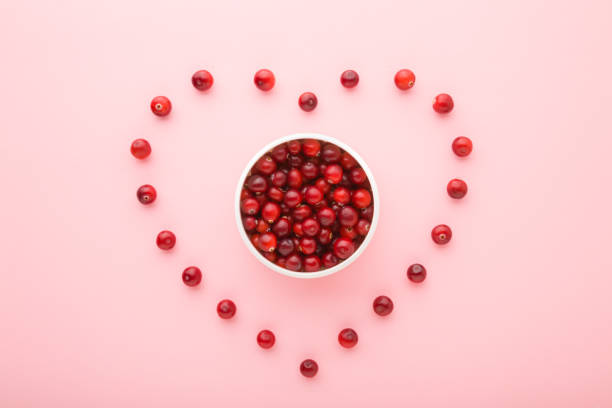 Heart shape created from red berries. Fresh cranberries in white bowl on light pink table background. Pastel color. Love healthy food. Closeup. Top down view. stock photo