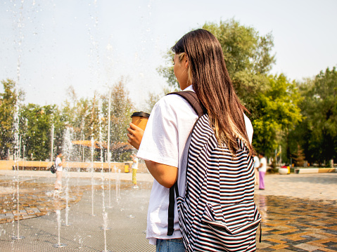 pedestrian fountain, young woman with a backpack walks next to the jets of an extensive water mirror fountain, Pleasant refreshment, relaxation in the summer heat, a walk in the summer park near the fountain