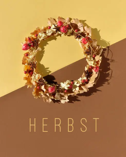 Dried floral wreath made of dry Autumn leaves and berries. Word Herbst means Autumn in German language. Seasonal Fall flat lay on brown and yellow paper background. Direct sunlight with shadows.