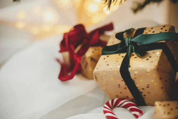 Photo of Merry Christmas! Stylish christmas gifts under christmas tree with golden lights. Wrapped christmas presents in golden paper with red and green ribbons in festive atmospheric room