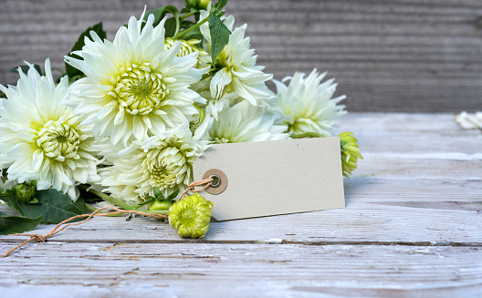 A bouquet of white dahlia flowers and a card with copy space are on a wooden background.