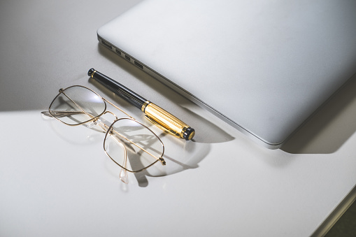 Minimalistic office, closed silver laptop, gold eyeglasses and gold pen.