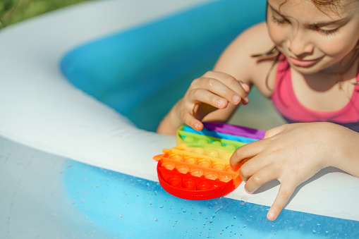 Little girl, kid, child plays, swims with colorful pop it. Funny antistress colorful sensory push toy popit in inflatable summer rubber swimming pool. Staycation at home in backyard of country house.