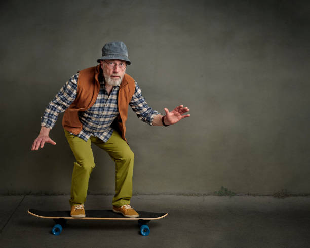 senior man riding a long cruising skateboard bearded senior man wearing round glasses and a bucket hat is riding a long skateboard in a grunge urban environment man beard plaid shirt stock pictures, royalty-free photos & images