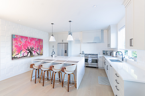 Big white spacious kitchen with a marble peninsula with wood bar stools, stainless steel stove and fridge and wall art and two hanging Pendant lights