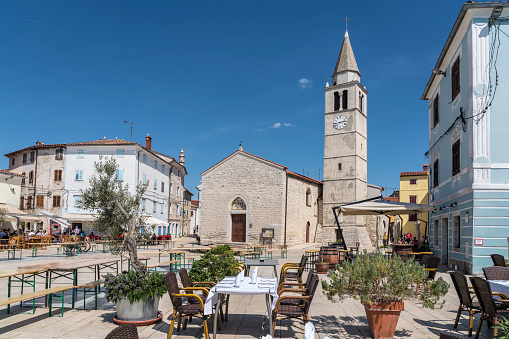 Some tourists on the main square in Adriatic city Fažana, from where the ferry goes to island Brijuni. In front empty tables set for lunch, in background small church.