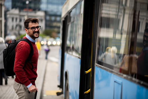 Handsome young man on his way to work through the city center, living the modern urban lifestyle. Attractive young businessman happy and smiling while boarding the bus. Fashionable young entrepreneur with his backpack getting on a bus.