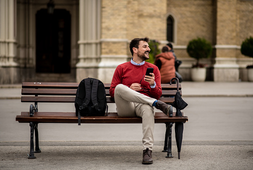 Handsome young man on his way to work through the city center, living the modern urban lifestyle. Attractive young businessman having a break, sitting on a bench. Fashionable young entrepreneur with his backpack texting on his phone.