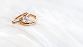 Pair of golden wedding rings on white textile background with copy space. Engagement ring with diamond on fabric backdrop. Symbol of love and marriage.