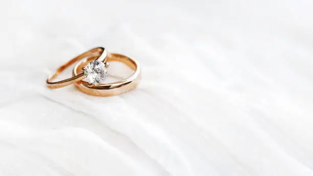 Photo of Pair of golden wedding rings on white textile background with copy space. Engagement ring with diamond on fabric backdrop. Symbol of love and marriage.