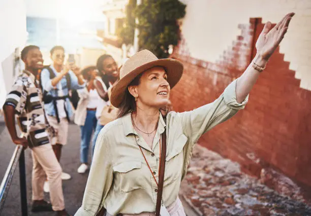 Photo of Travel, education and a teacher with students on school field trip, on urban tour. Woman, city guide and group of happy tourists, pointing at local architecture and learning on international holiday.