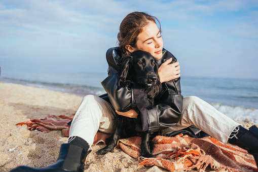 Portrait happy serene young woman hugging her dog while sitting on plaid on sandy sea coast on sunny day