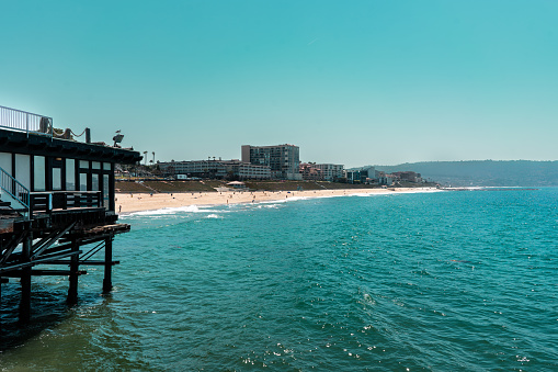 Redondo Beach, CA, USA - August 2016: An ocean view from a pier with clear blue skies. A building above the water can be seen on the left and in the distance, people can be seen on the sand along the coast. Tall buildings can be seen in the distance.