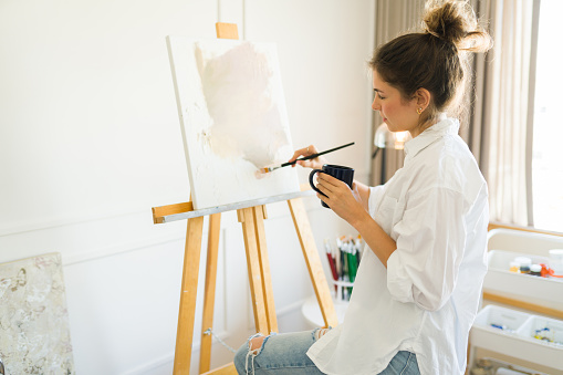 Caucasian woman in her 20s enjoying her art hobby drinking coffee while painting on a canvas with an easel