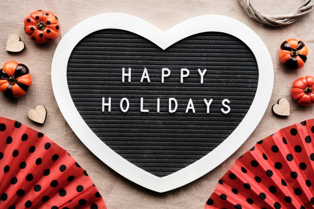 Halloween orange black flat lay background with decorations. Heart shaped felt letter board with text Happy Holidays. Orange fans with polka dots, painted mini deco pumpkins with black stripes and circles.
