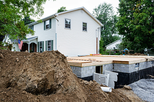An in-process suburban residential home improvement in-law apartment addition construction project. The pile of dirt and debris in the foreground was excavated from the deep hole dug for the new basement foundation. Progress has been made on installation of the mandatory egress/escape hatch door/window required by local town building codes and fire evacuation safety regulations.
