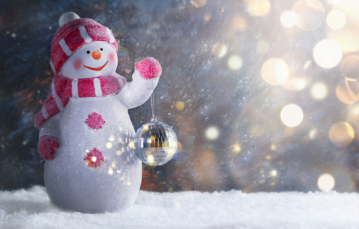 Cute happy snowman with Christmas ornament on a snowy winter night. Christmas background. New Year greeting card. Fabulous Christmas composition with snowman dressed in warm clothes, mittens, scarf and hat.