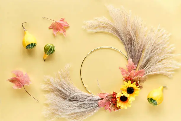 Autumn background on yellow paper. Dried floral wreath and deco gourds. Dry pampas grass, flowers and Autumn leaves on metal frame. Flat lay on white yellow table. Soft natural sunlight.