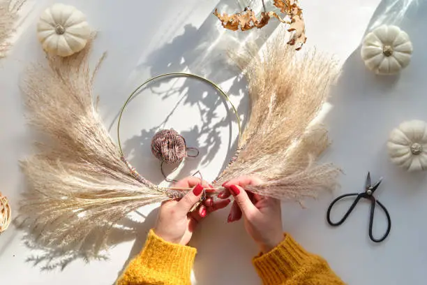 Hands making dried floral wreath from dry pampas grass and Autumn leaves. Hands in sweater with manicured nails tie decorations to metal frame. Flat lay on white table, sunlight with long shadows.