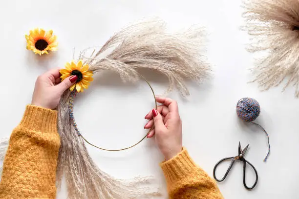 Hands making dried floral wreath from dry pampas grass and Autumn leaves. Hands in sweater with manicured nails tie decorations to metal frame. Flat lay on white table, sunlight with long shadows.