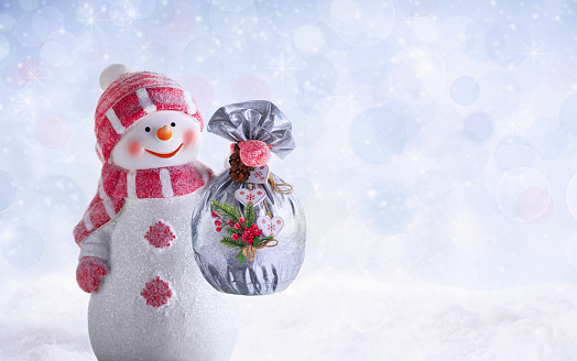 Happy snowman holds a bag with Christmas presents, dressed in warm clothes, mittens, hat and scarf in a frosty winter day. Winter or Christmas background or banner with defocused lights and lens flare.