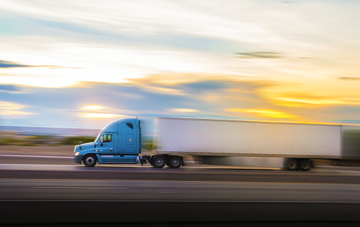 Blue and white semi-truck driving on the highway at sunset