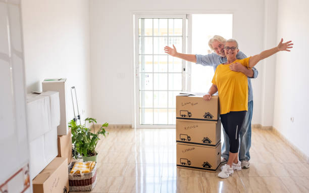 istock Smiling caucasian senior couple white haired open arms looking at camera during relocation, happy for the new beginning. Moving boxes on the floor, empty apartment 1431254981