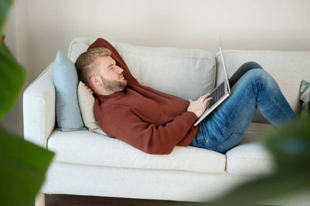 istock Young caucasian blond curly hair man lying on sofa looking at laptop, using computer,working online at home in modern scandinavian interior 1431261807