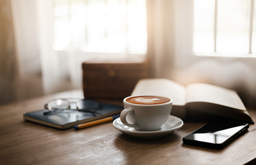 Close up view, Latte coffee in white cup and smart phone on wooden table near bright window. blurred background with book, eyeglasses on blue note book and pen, sepia color tone